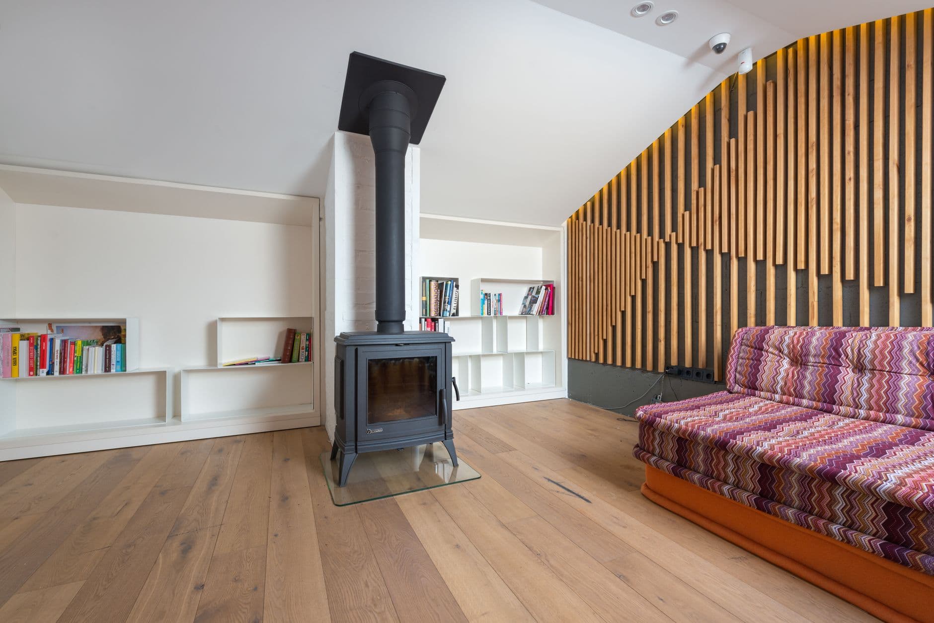 Comfortable sofa placed near wall decorated with wooden planks and fireplace against shelves with books